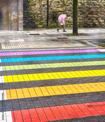 Rainbow crosswalk during a rainy day. A vibrant rainbow painted crosswalk on a wet city street, with a person walking in the background under a pink umbrella during Pride month celebrations