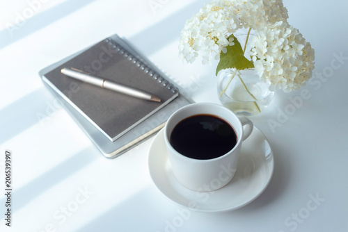 Coffee cup, notebook and white hydrangea flowers on a bright desk with copy space