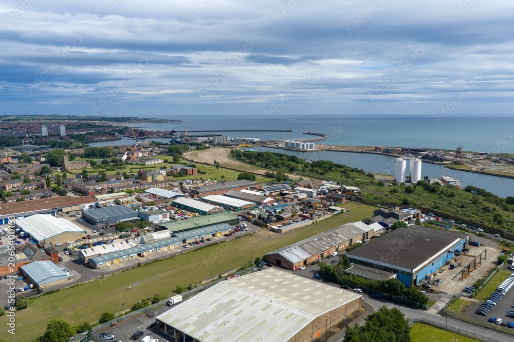 Fototapeta premium Aerial view across the Port of Sunderland showing harbour entrances industrial facilities warehouses riverside infrastructure and the North Sea coastline beneath dramatic cloud formations summer day