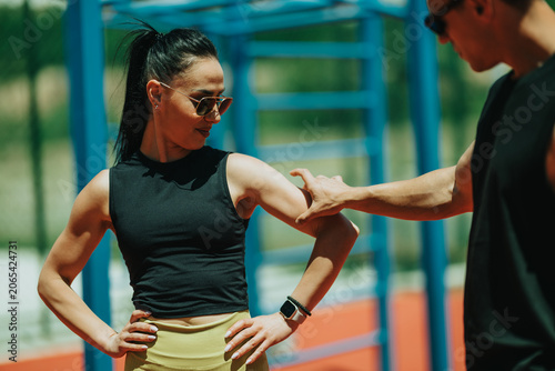 Female athlete listens to a coach while stretching her arm during an outdoor training session. The coach provides hands-on guidance during a sunny fitness workout on a sports court.