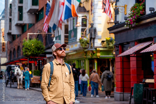 Fotografie A man stands in the iconic Temple Bar district of Dublin, looking up amidst the characteristic flags and bustling atmosphere of the famous street