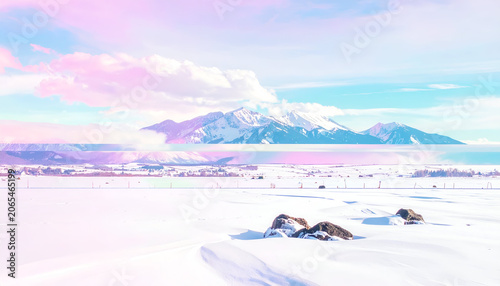 Snowy winter scene with pastel skies, mountains, clouds, & snow-covered foreground rocks