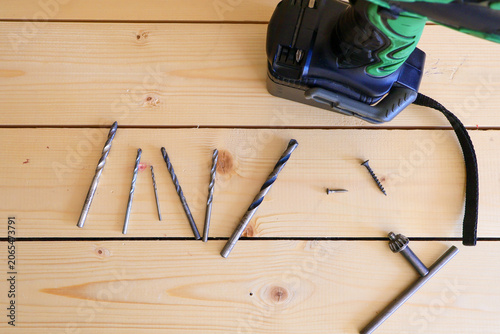Cordless drill with green accents rests on light wood planks. Scattered drill bits, screws, and a hand brace convey preparation for hands-on construction work.