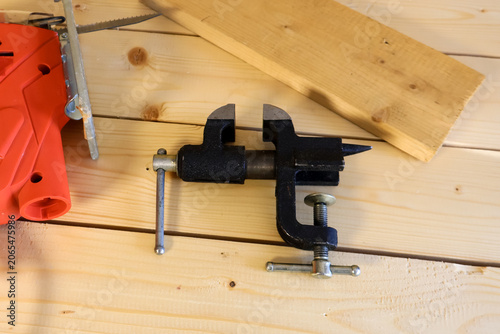 Compact black bench vise with T-handle and screw mechanism sits on light pine planks. Nearby, a wooden board and partial view of a red power tool convey a prepared, hands-on workspace.