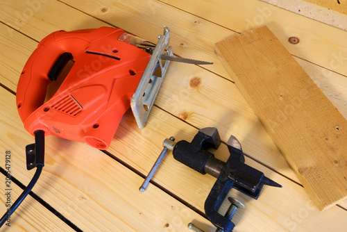 Bright orange electric jigsaw sits on light pine planks with blade exposed. Black bench clamp and raw pine board signal preparation for accurate woodworking tasks