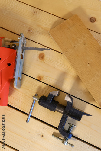 Metal hand saw with sharp teeth rests near a black bench clamp with silver hardware. Light pine board and wooden planks create a clean, ready-to-work atmosphere for hands-on carpentry