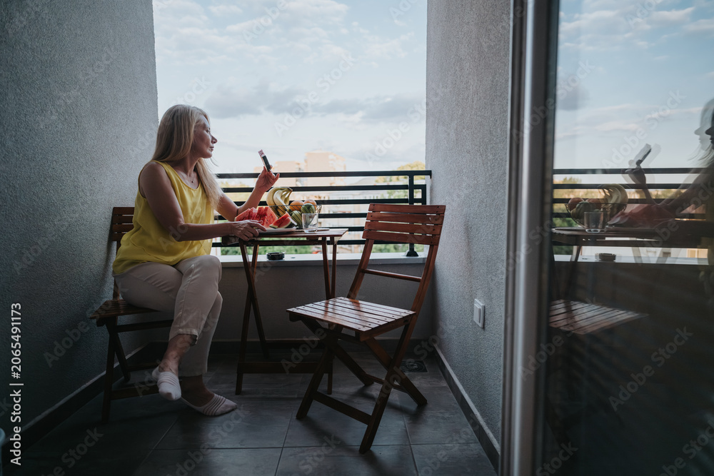 Fototapeta premium A mature woman sits on a small balcony holding a smartphone beside a plate of fresh fruit. She relaxes on wooden chairs while checking her phone and enjoying the view.