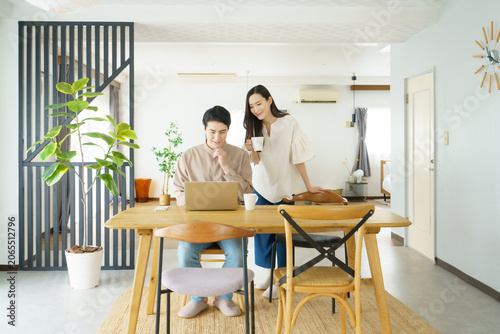 自宅のテーブルでノートパソコンで作業をする男性とマグカップを持つ女性の若い日本人パワーカップル