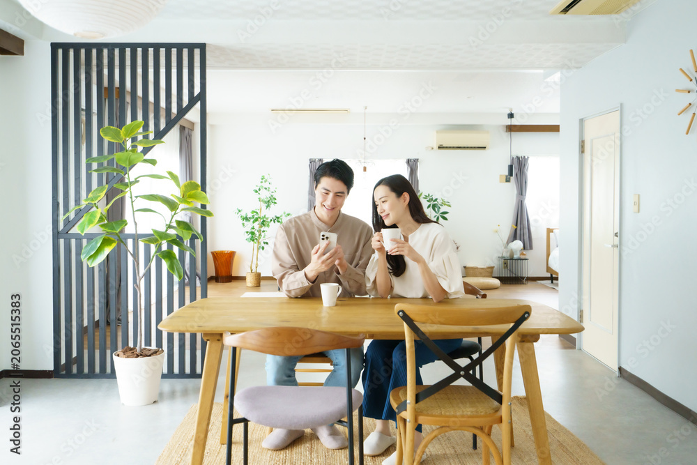 Fototapeta premium 自宅のテーブルでスマホを見ながら飲み物を飲む若い日本人パワーカップル