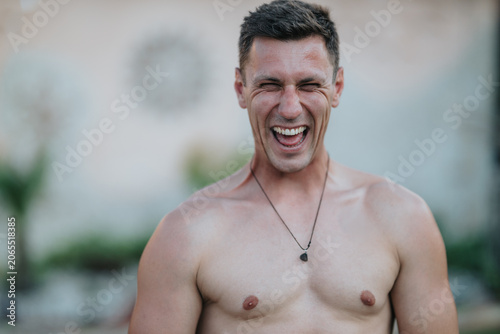 Shirtless young man laughing with eyes closed showing a wide smile and yin yang tattoo. Casual outdoor portrait capturing joyful, candid emotion and relaxed energy.