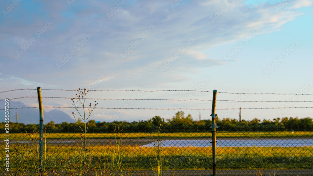Fototapeta premium Wire mesh fence with barbed top overlooking a field and distant mountains