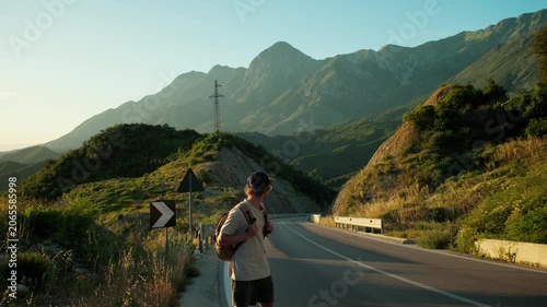 Handheld wide shot of male traveler standing on mountain road, scanning for cars and extending arm to hitchhike. Backpack, roadside curve and sunset peaks frame waiting and travel uncertainty.