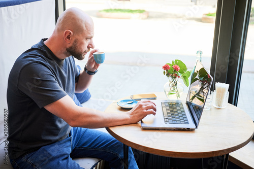 Man working on laptop and drinking espresso in cafe, remote work by window with coffee, flowers and water on table, freelance lifestyle and productivity concept