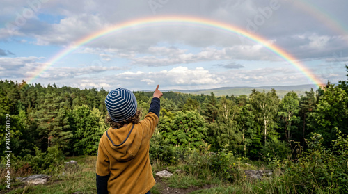 A child in a yellow jacket seen from behind points at a wide rainbow curving over a lush green forest canopy from a hilltop viewpoint under partly cloudy skies, an evocative image for nature and outdo