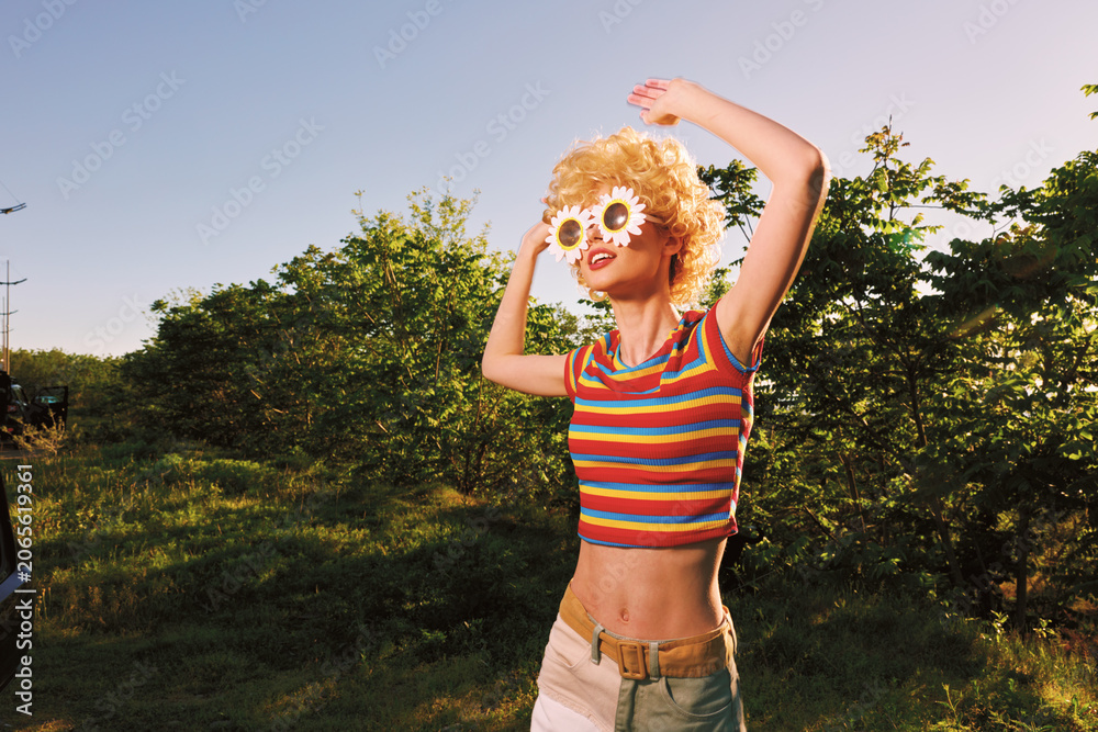 Obraz premium Woman enjoying sunny outdoor moment in retro film color, wearing striped crop top and light shorts, flower sunglasses, dancing arms up, carefree lifestyle, vibrant natural setting