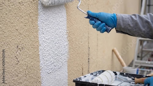 Handheld tracking: painter's gloved hand rolls white paint on textured stucco exterior; close-up of roller and surface; ladder and scaffold visible.