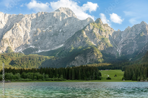 Panoramic view of Laghi di Fusine in Italy with a crystal-clear lake, forest and majestic Julian Alps under blue sky. Scenic alpine landscape for travel, nature and outdoor adventure themes.