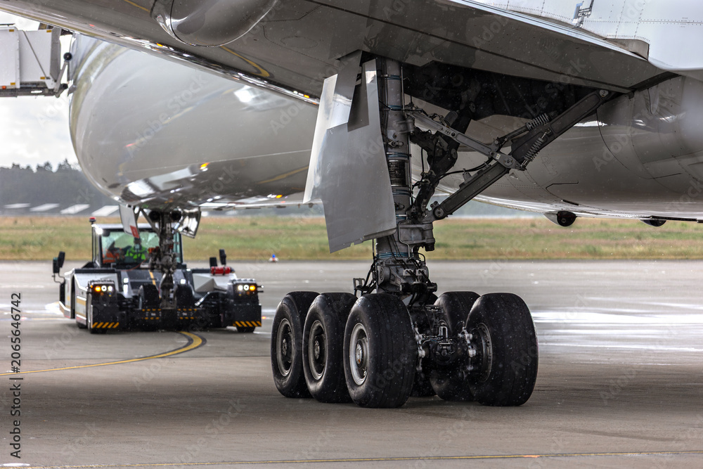 Fototapeta premium Widebody aircraft undergoes pushback operation wet airport apron heavy tow tractor maneuvers fuselage. Detail landing gear assembly rain aviation engineering ground support operational efficiency