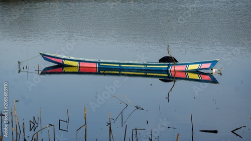Canvastavla A vibrant, colorfully painted traditional wooden boat floats on the calm, mirror-like surface of a serene lake during a clear day