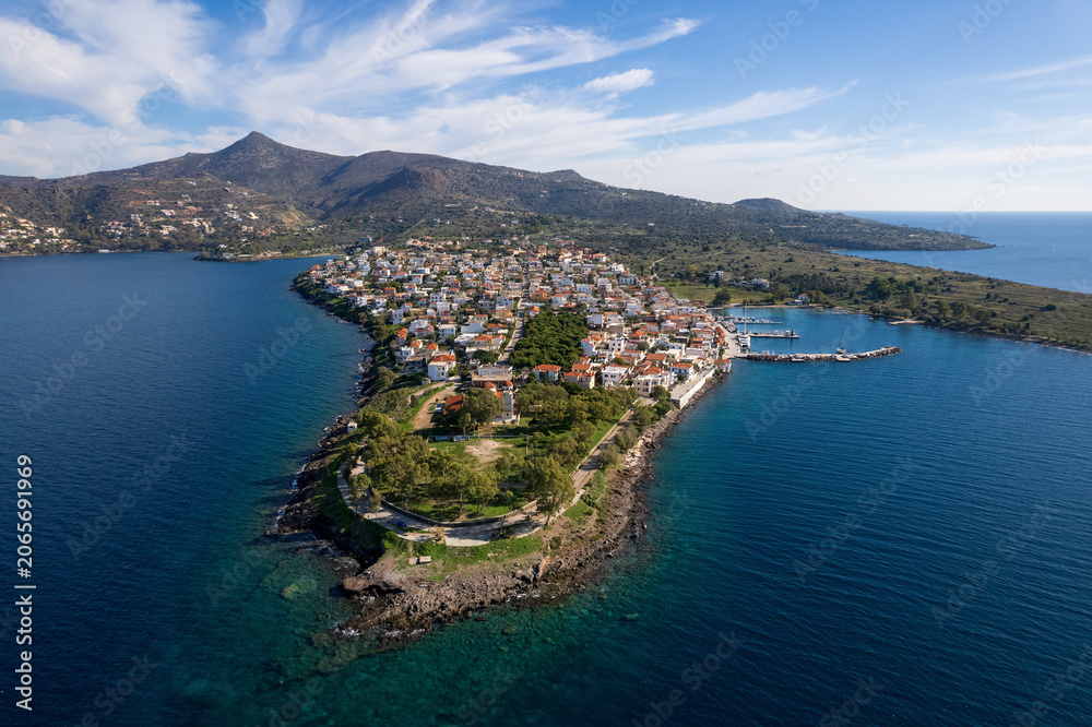 Fototapeta premium Drone shot of the traditional coastal village of Perdika on Aegina island. White houses and a scenic marina stretch along a rocky peninsula into the deep blue Saronic sea, with mountains in background