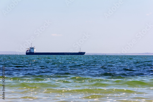 Long cargo ship navigates calm ocean. Water shimmers with green, yellow, deep blue hues. Clear light blue sky dominates upper frame, distant land visible