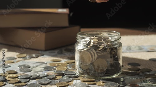 Hand Adding Coin Into Savings Jar With Cash and Books Background, budgeting strategy for education and future expenses