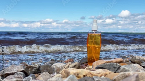 Tanning lotion bottle stands on pebble beach while lake waves roll in the background under bright summer sky.