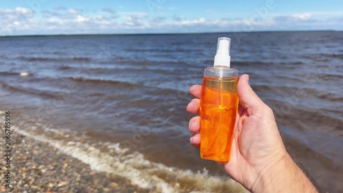 Male hand holds tanning lotion spray bottle near lake shore in sunny summer weather.