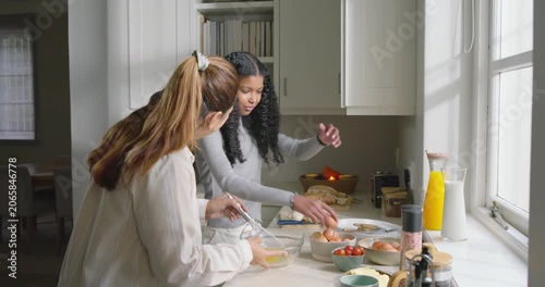 African American mom and daughter cracking eggs and whisking at counter by window making breakfast