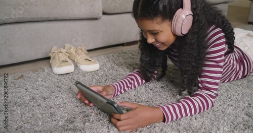 Teen African American girl wearing striped top lying on rug at home with headphones holding tablet