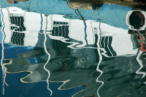 Fotografie An abstract view captures the wavy and distorted mirror reflection of a white boat hull with black windows on the water surface