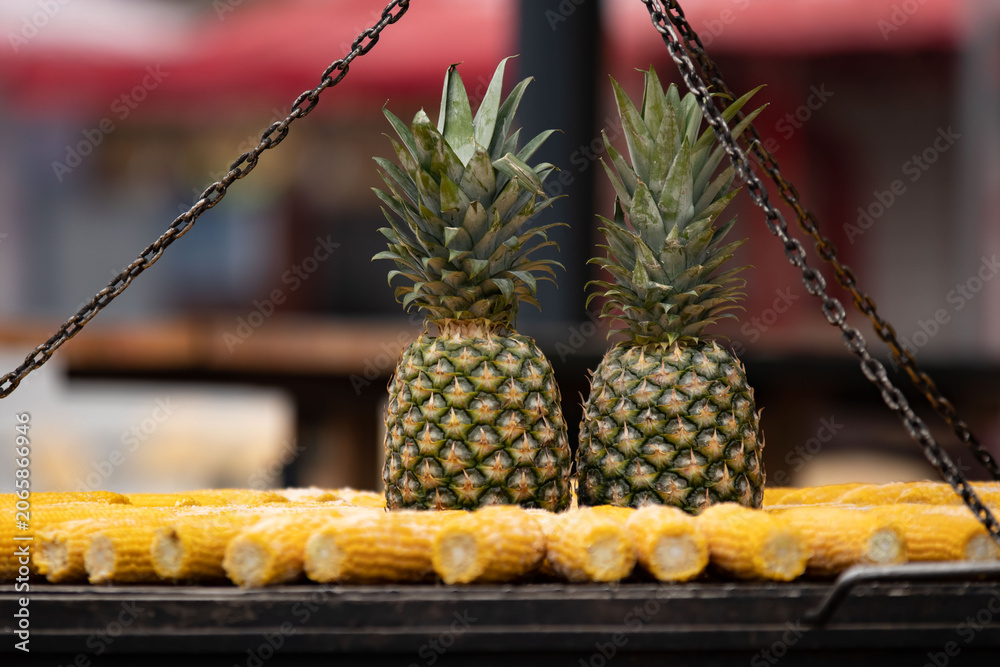 Obraz premium Eye-level shot of two whole pineapples sitting on iron grill rack with corn cobs below, metal chains frame both sides. Blurred red market awning behind creates vivid urban street food atmosphere