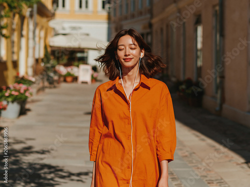 Short-haired East Asian lady in loose dusty pink linen shirt closes eyes smiling blissfully with wired white earbuds, walking sun-dappled old European street lined with flower terracotta pots.