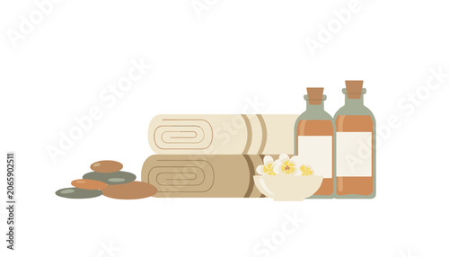 Stacked soft towels beside a bowl of white flowers, massage stones, and amber glass bottles containing essential oils for spa therapy.