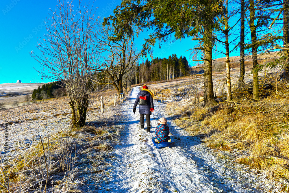 Fototapeta premium Mutter zieht ihr Kind auf dem Schlitten bei einer Winterwanderung auf der Wasserkuppe in der Rhön, Hessen