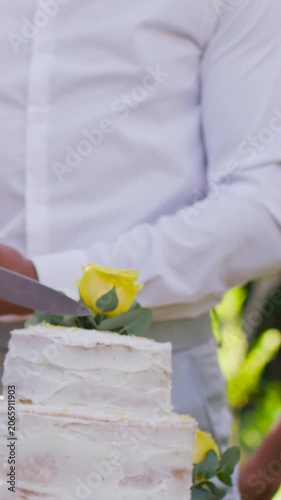 Vertical video: Lowering knife man white shirt slicing tiered cake with yellow roses at yard party