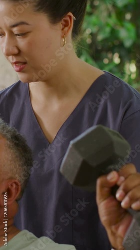 Vertical video: Supporting therapist in scrubs steadying patient starting hex dumbbell press patio