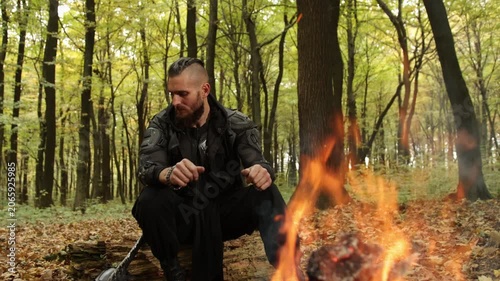 Campfire warmth, hands warming, wilderness fire. Survivalist dressed in tactical clothing, with braids in his hair, warms his hands near campfire in heavily wooded area.