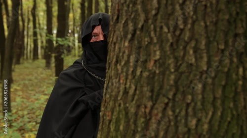 Secrecy suspense, Shadowy presence, Hand grasp. Gripping tree trunk in shadowy forest, hooded figure with painted hand evokes secrecy and suspense.
