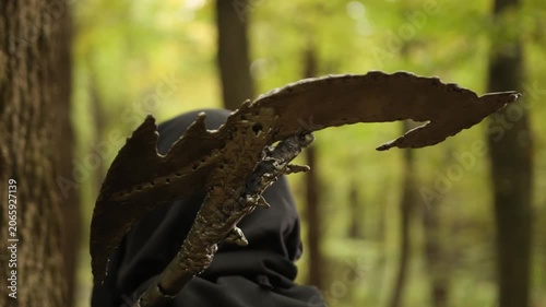 Weapon detail, Weapon closeup, Forest ambiance. Hooded person clutching rugged, self-made weapon, set before softly blurred forest scene.