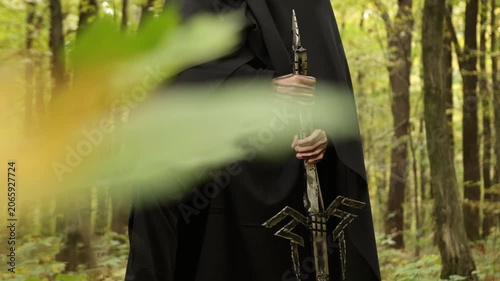 Cloaked figure, woodland setting, forest mood. Close-up cloaked figure in black holding intricately designed sword, set against forest background with blurred leaves.