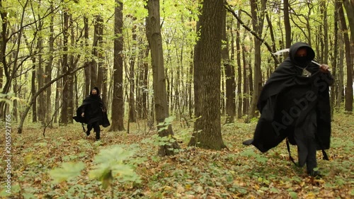 enemy's attack, forest path, occult forest. Two hooded individuals dressed in dark robes walk through thick autumn woodland, creating mood filled with fantasy or horror and intense suspense.
