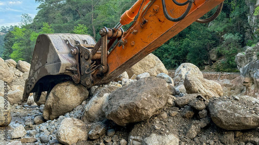 Fototapeta premium Excavator bucket lifting boulders in a forest riverbed at soft daylight, ideal for construction editorial, environmental restoration, infrastructure use, 5 May 2026 Subitica, Serbia
