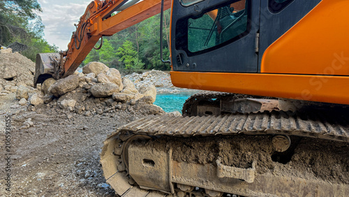 Orange excavator on rocky riverbank in soft daylight by turquoise water ideal for construction editorial environmental reports and social media, 5 May 2026 Subitica, Serbia