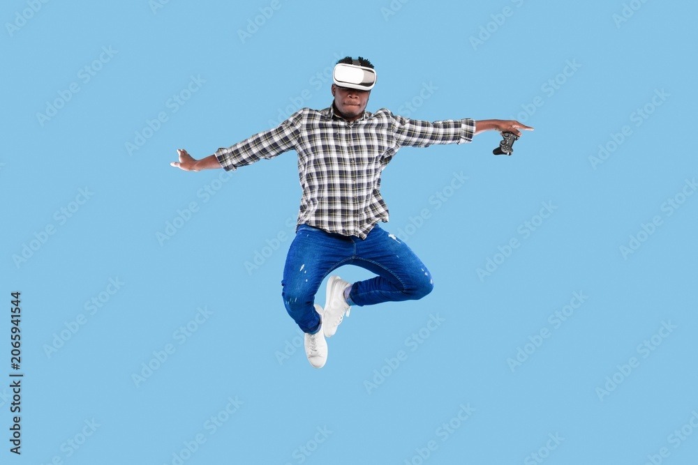 Obraz premium Excited Young Black Man Engaging With Virtual Reality Headset While Jumping In Indoor Studio Setting, Holding Game Controller. Enjoying New Gaming Experience In Modern Environment.