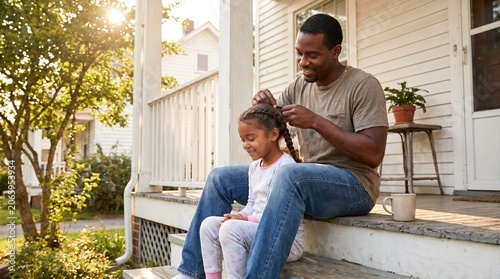 A father braiding his daughter's hair on their porch at sunset, creating a heartwarming moment of connection