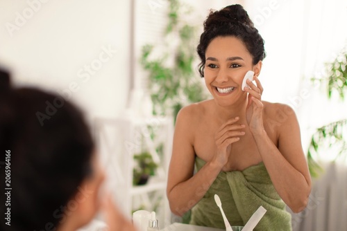 Konstfotografi Smiling Young Woman With Olive Skin Applying Facial Cream In Front Of Mirror At Home, Taking Care Of Her Skin With A Daily Routine In A Bright, Relaxing Setting