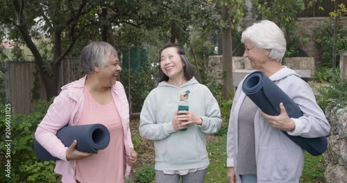 Diverse female friends arriving in garden, greeting, holding yoga mats and teal bottle for yoga