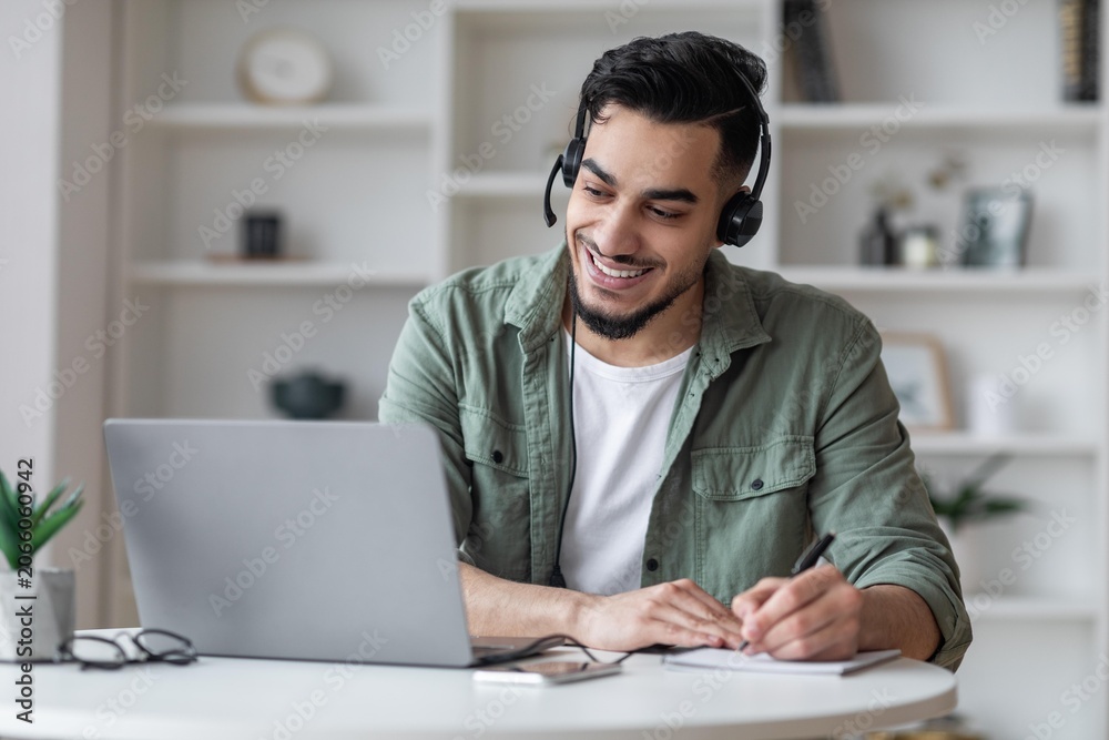 Obraz premium Smiling Young Hispanic Man Engaged In Remote Work With Laptop And Headphones In Cozy Home Office Setting. Focused On Tasks, Taking Notes, Representing Modern Business Concept.