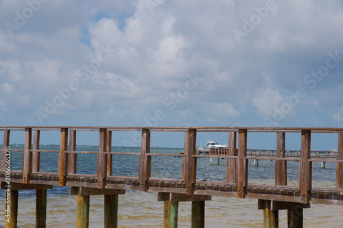 Foto A rustic wooden pier stretches across calm coastal water toward a docked boat under a broad cloudy blue sky, evoking summer relaxation, seaside travel and peaceful waterfront scenery